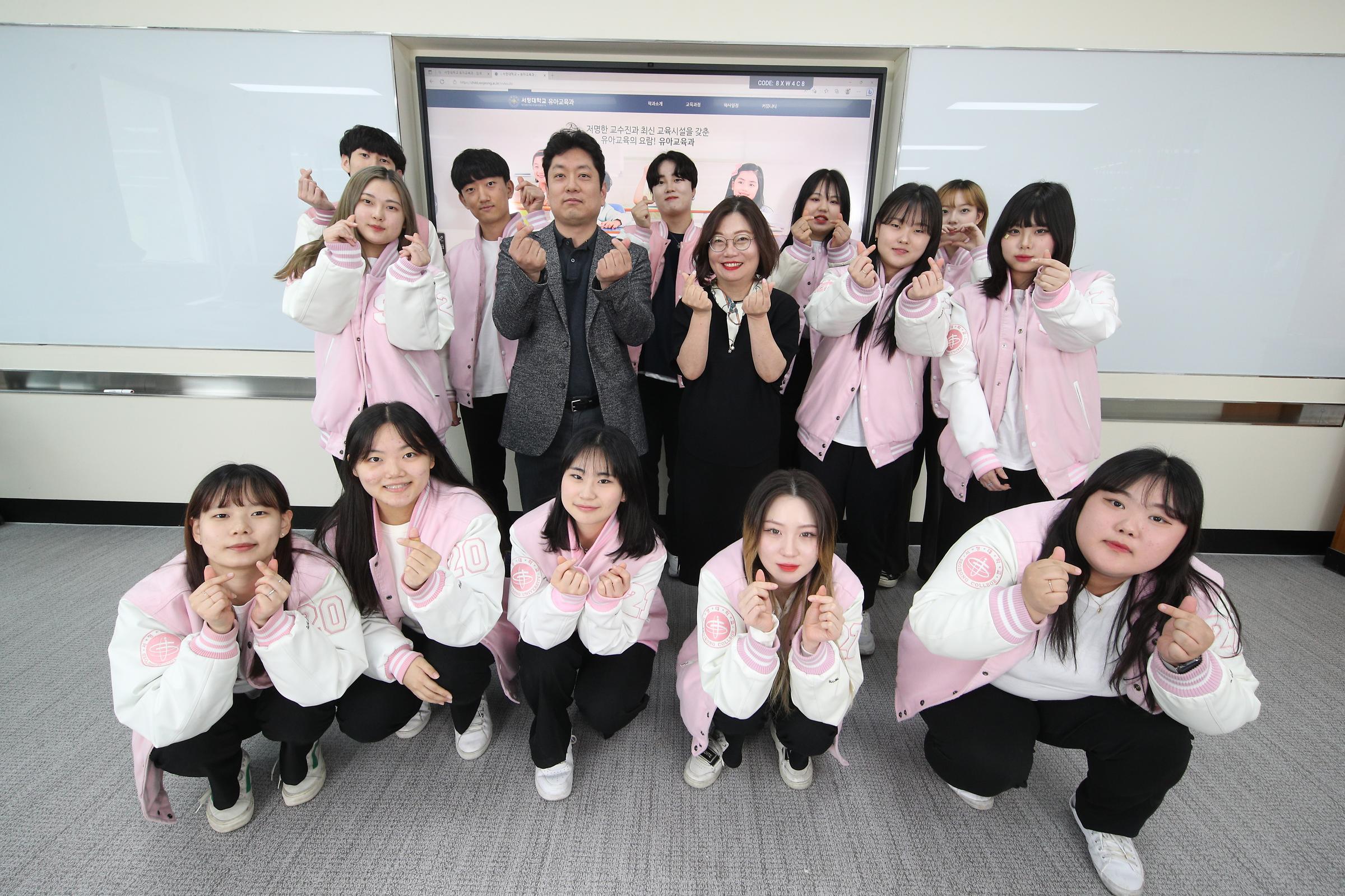 분홍색 과 점퍼를 입고 모여있는 간호학과 학생들과 가운데 서있는 두 명의 교수님들 사진.모두 웃으며 손가락 하트를 만들어 보이고 잇다.